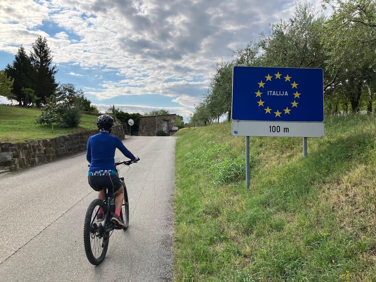 Cyclist approaching EU border sign for Italija (Italy) on a paved country road.