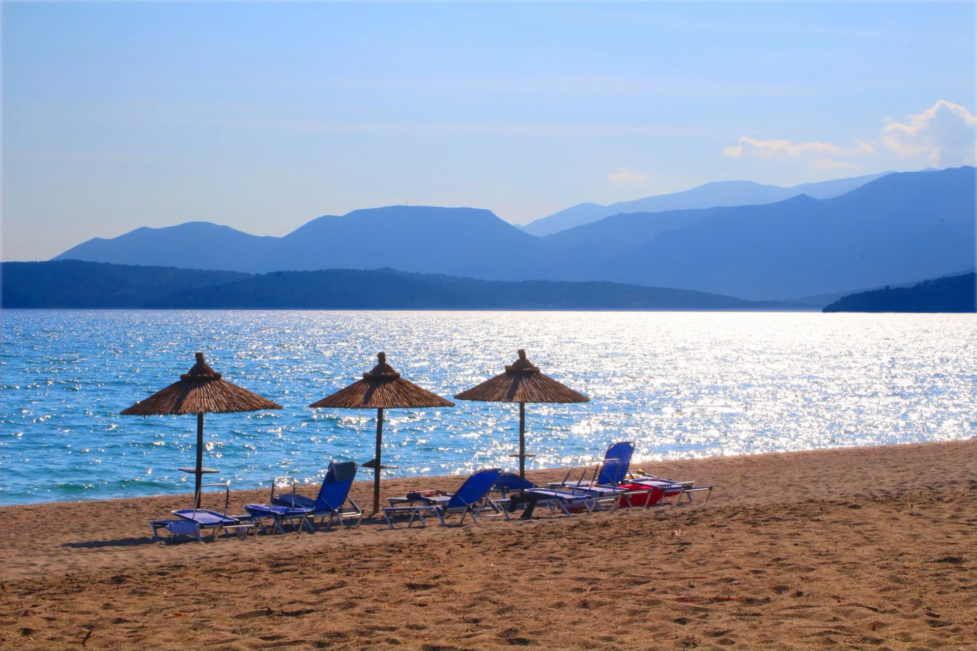 Beach chairs and straw umbrellas on sand facing sparkling blue water and blue mountains.