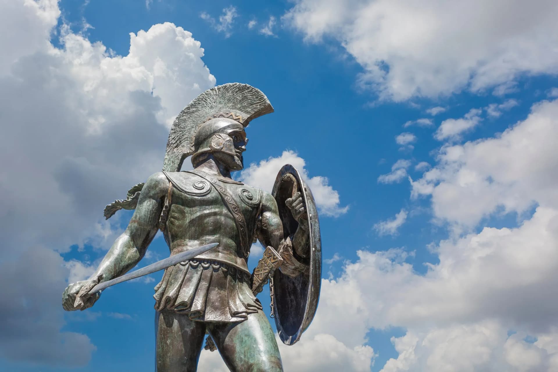 Bronze statue of a Spartan warrior with a helmet, sword, and shield against a bright blue sky with white clouds.
