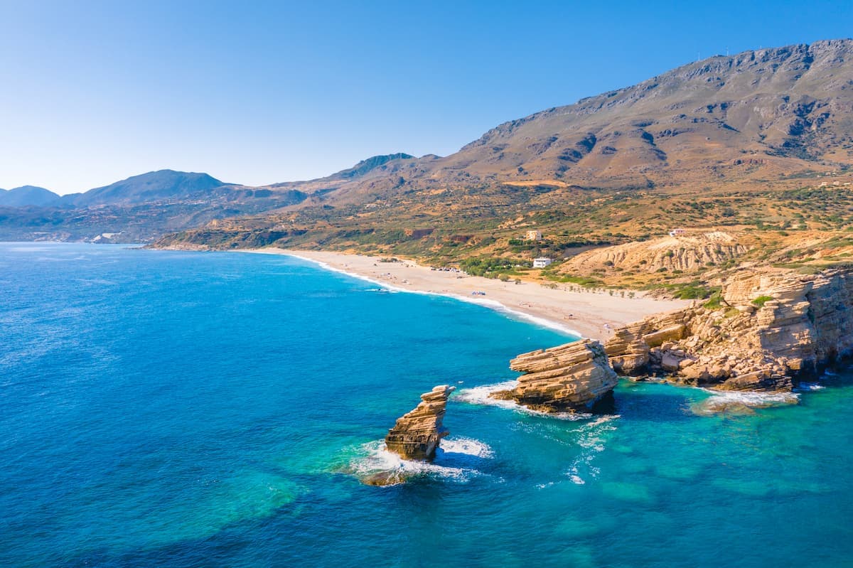 Coastal beach with turquoise water, rock formations, and arid mountains under a clear blue sky.