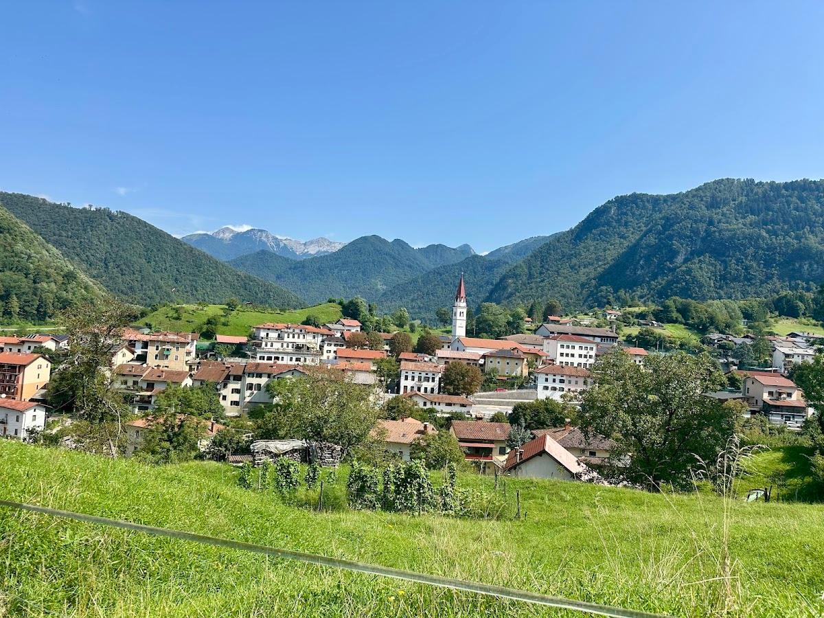 Village nestled in green valley with church tower, backed by forested mountains and snowy peaks.