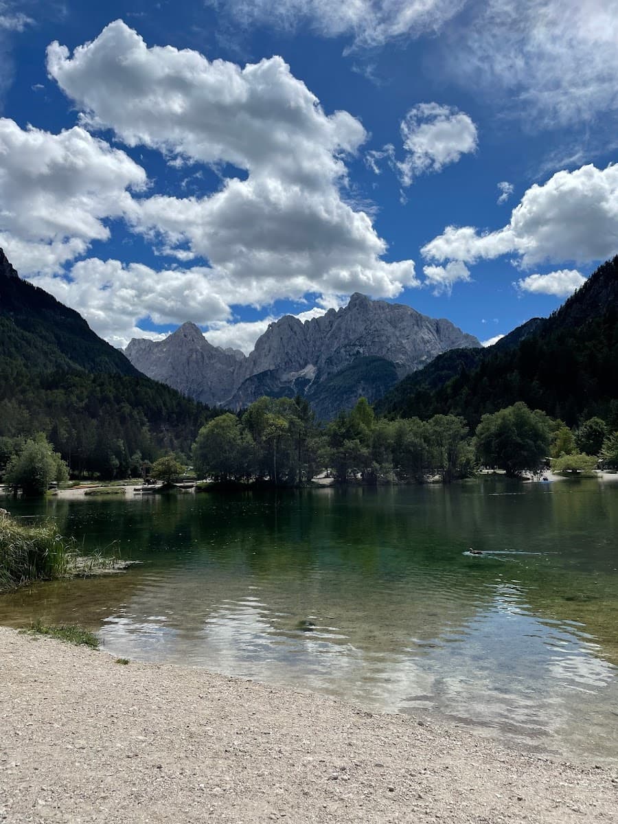 Mountain lake with clear water, forested shore, and jagged peaks under a blue cloudy sky
