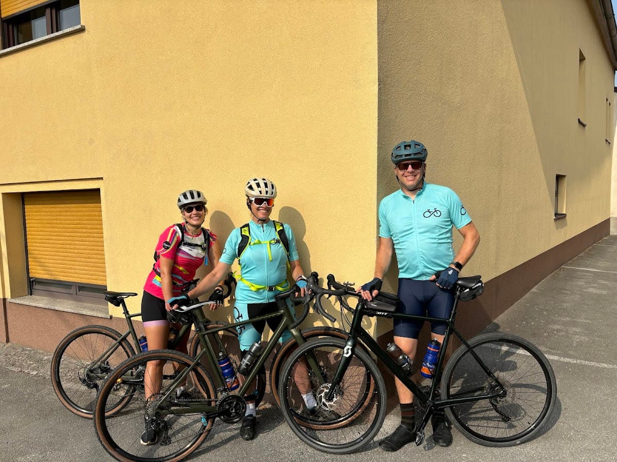 Three cyclists with gravel bikes posing outdoors against a yellow stucco building wall.