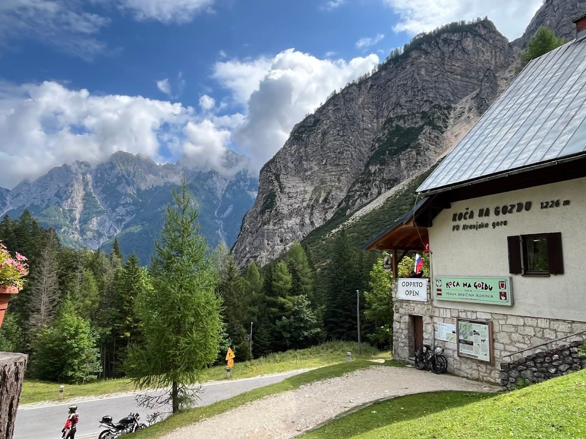 Mountain hut Koča na Gozdu with motorcycles parked near a road against a backdrop of steep, forested mountains.